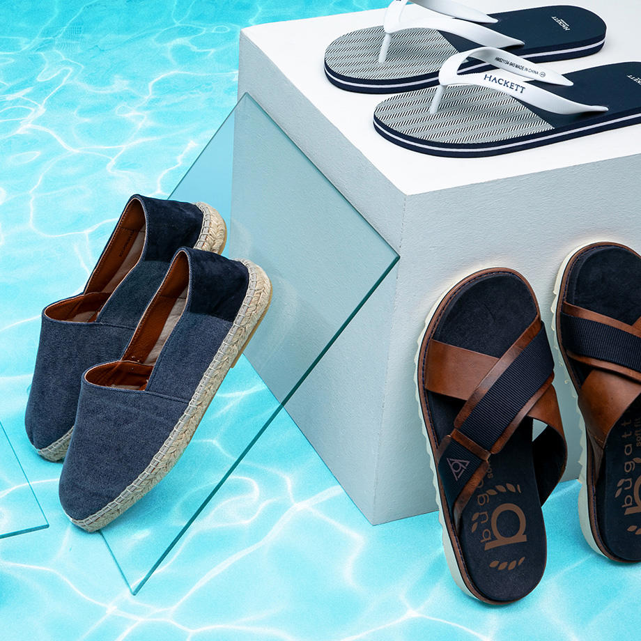 Verschiedene Sommerschuhe, darunter Espadrilles und Zehentrenner in Blau und Weiß, arrangiert am Poolrand auf hellen Podesten mit Wasser im Hintergrund.