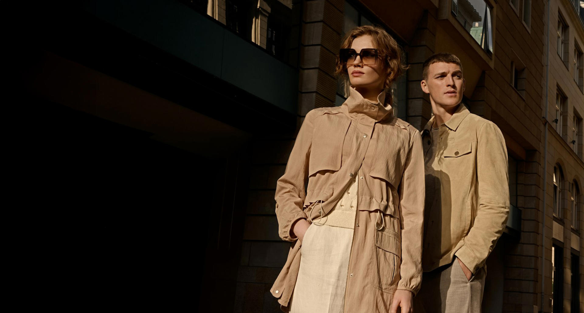 Ein Mann und eine Frau stehen in einer Stadt mit einem Gebäude im Hintergrund. Sie beide tragen beige Looks.