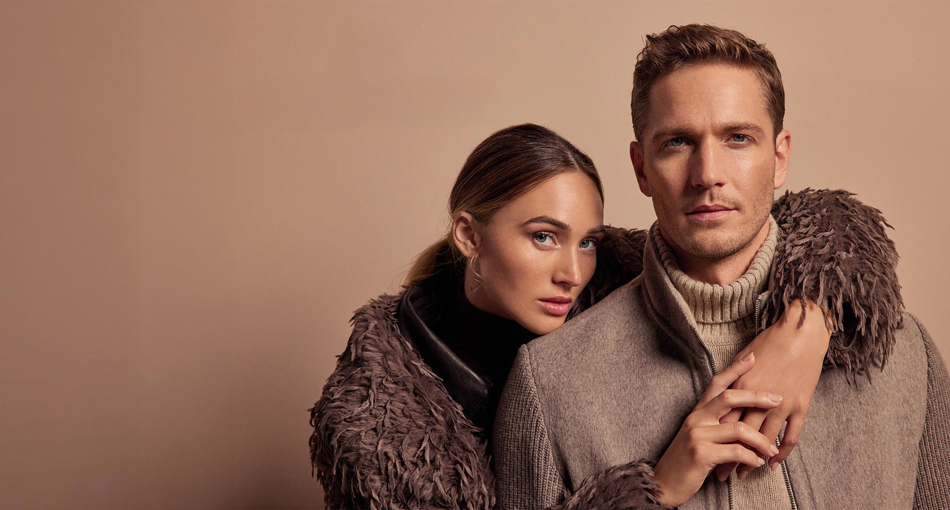 Ein Mann und eine Frau stehen in einer Stadt mit einem Gebäude im Hintergrund. Sie beide tragen beige Looks.