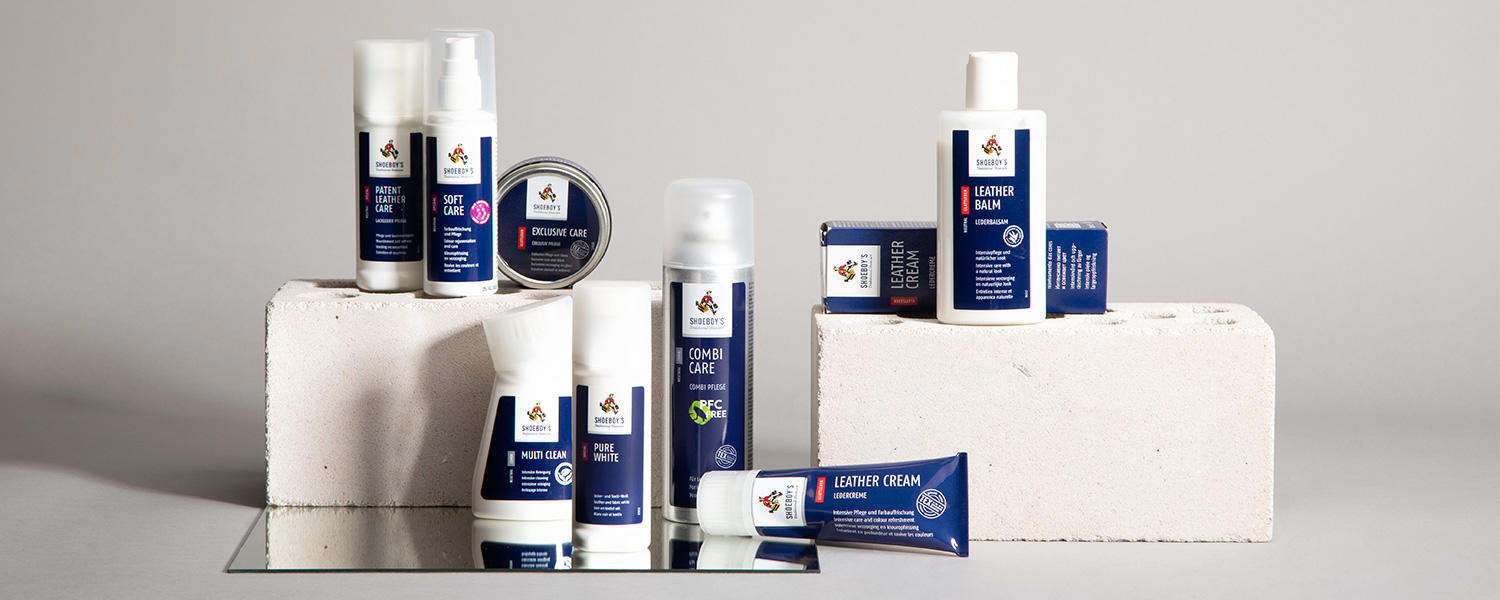 Pflegeprodukte von Shoeboy's, darunter Sprays, Tuben und Cremes, ordentlich arrangiert auf weißen Blöcken vor neutralem Hintergrund.
