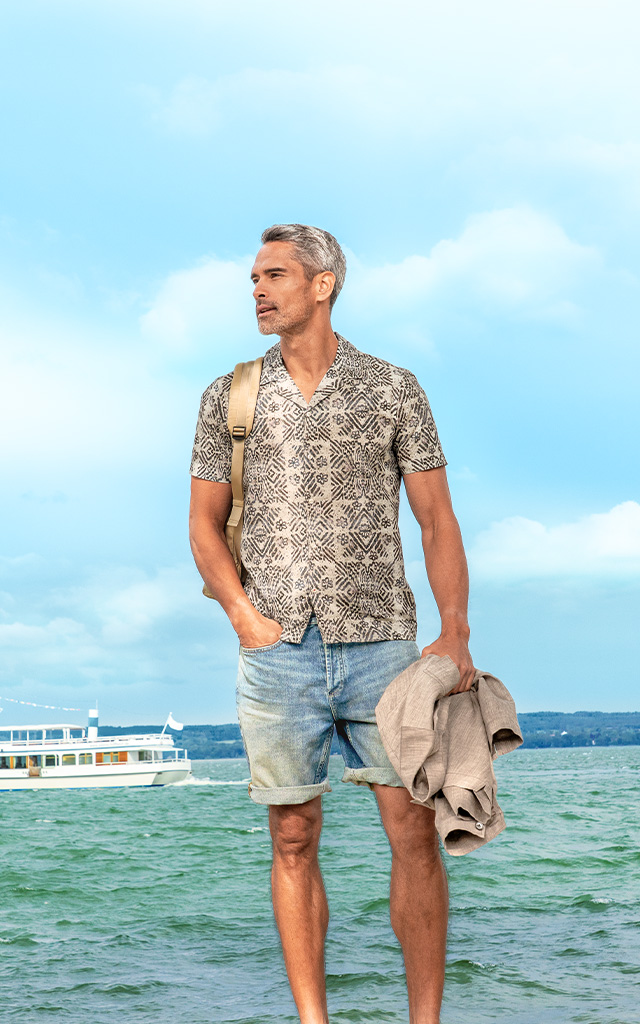 Ein Mann steht entspannt am Ufer eines Sees mit türkisfarbenem Wasser. Er trägt ein gemustertes Kurzarmhemd, Jeansshorts und hält eine Jacke in der Hand. Im Hintergrund fahren Segel- und Ausflugsschiffe.
