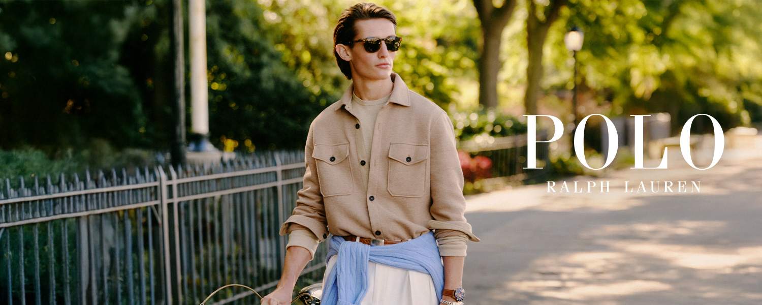 Ein Mann mit beiger Overshirt-Jacke, weißer Hose und braunen Loafern schiebt ein Fahrrad auf einem sonnigen Weg im Park. Großes POLO Ralph Lauren Logo im Vordergrund.