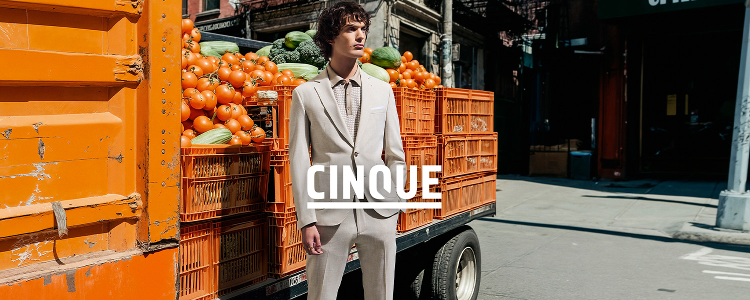 Ein Mann im beigefarbenen Anzug steht vor einem Lieferwagen voller Obstkisten. Aufschrift: 'CINQUE'