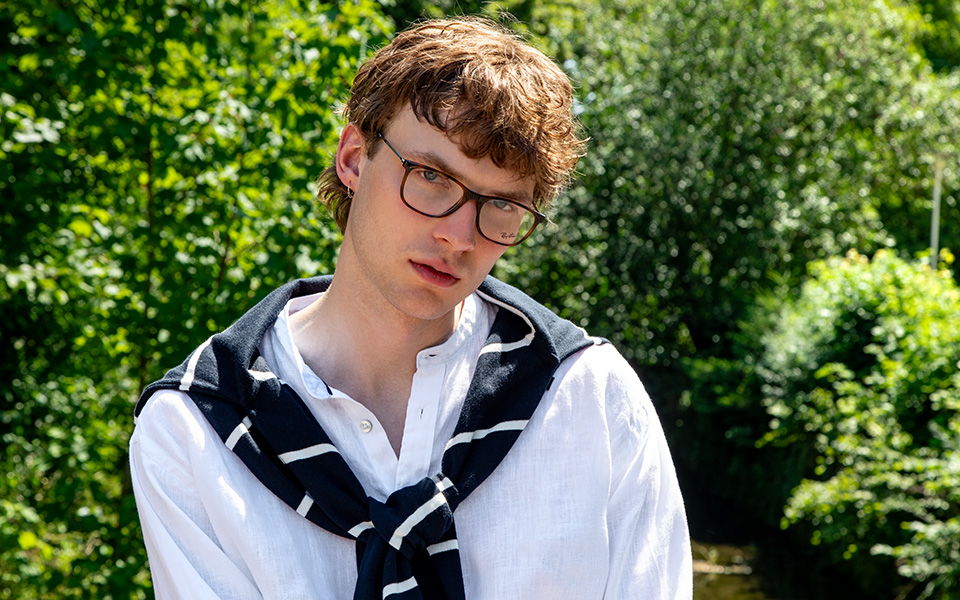 Ein junger Mann mit Brille vor einer grünen Szenerie. Er trägt ein weißes Leinenhemd und hat einen navy Strickpullover umgebunden.