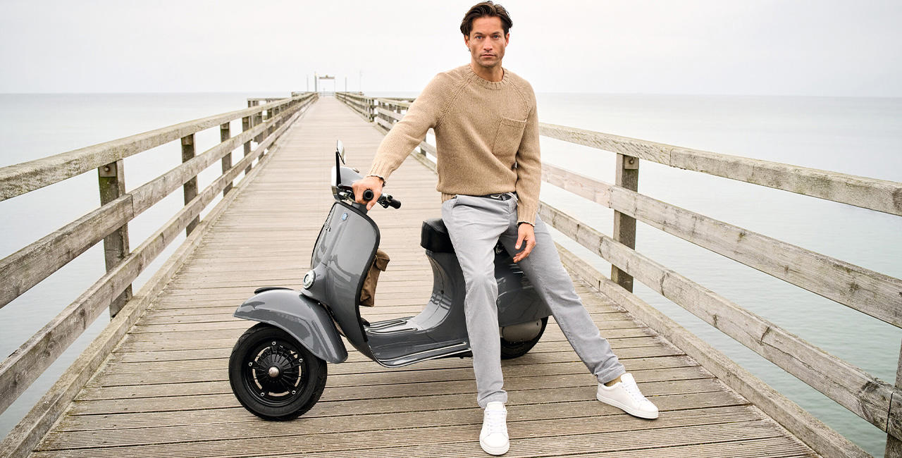 Mann in hellem Strickpullover und grauen Casual Pants lehnt entspannt an grauer Vespa auf Holzsteg am Meer – moderner Lifestyle-Look von Alberto.