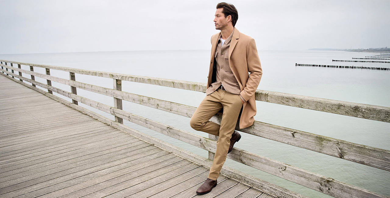Stilvoller Mann im beigen Hose, Overshirt und Mantel sitzt lässig auf einem Holzsteg am Meer – elegante Hosenmode für Business und Freizeit.