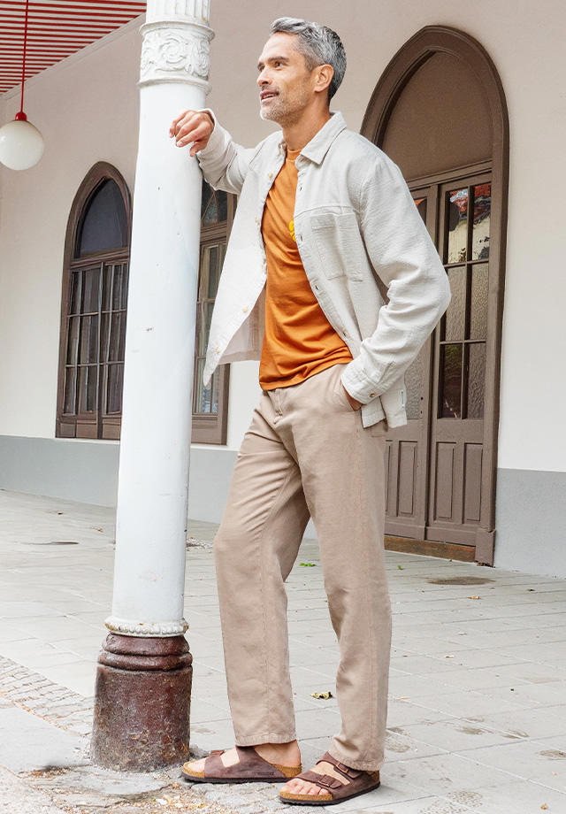 Stilvoll gekleideter Mann lehnt entspannt an Säule, er trägt ein beiges Overshirt und eine beige Hose mit Sandalen und orangefarbenem Shirt. 