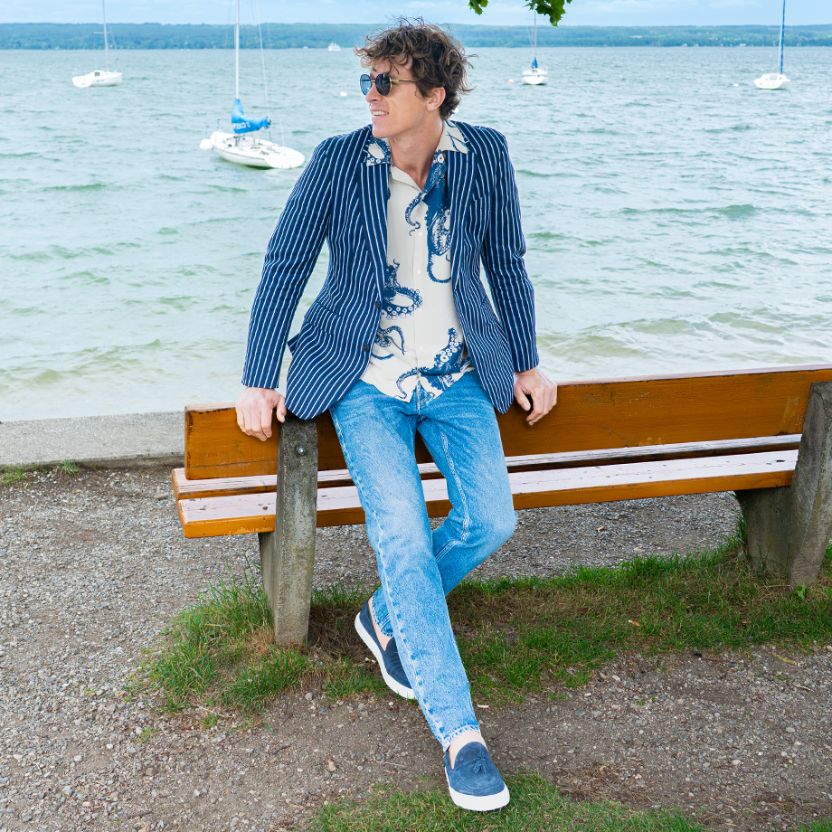 Mann in Jeans und gestreiftem Sakko sitzt entspannt auf einer Parkbank am See mit Segelbooten im Hintergrund.