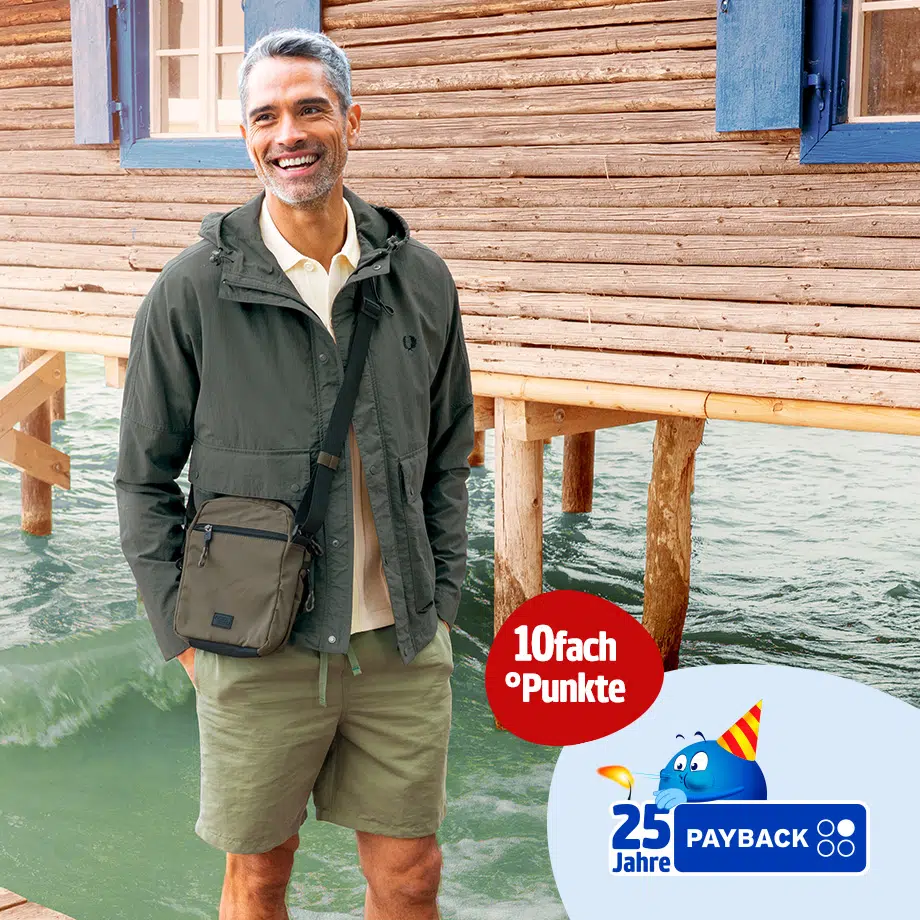 Lächelnder Mann in Outdoor-Look mit dunkelgrüner Jacke, Shorts und Umhängetasche steht vor einem Holzhaus am Wasser – Kampagnenmotiv mit Payback-Logo und Hinweis auf 10-fach Punkte zum 25-jährigen Jubiläum.
