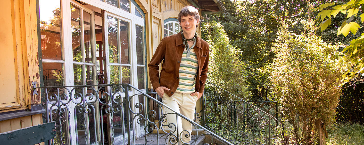 Ein junger Mann steht auf einer Treppe vor einem alten Haus. Er trägt ein Outfit in Herbstfarben: Braun, Grün, Beige.