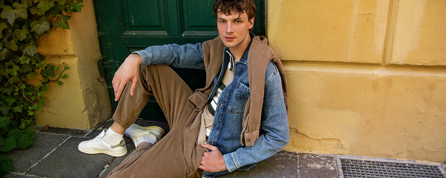 Ein junger Mann lehnt an einer alten Tür. Er trägt eine Cordhose, Poloshirt und eine Jeansjacke. Ein Cordhemd liegt über seinen Schultern