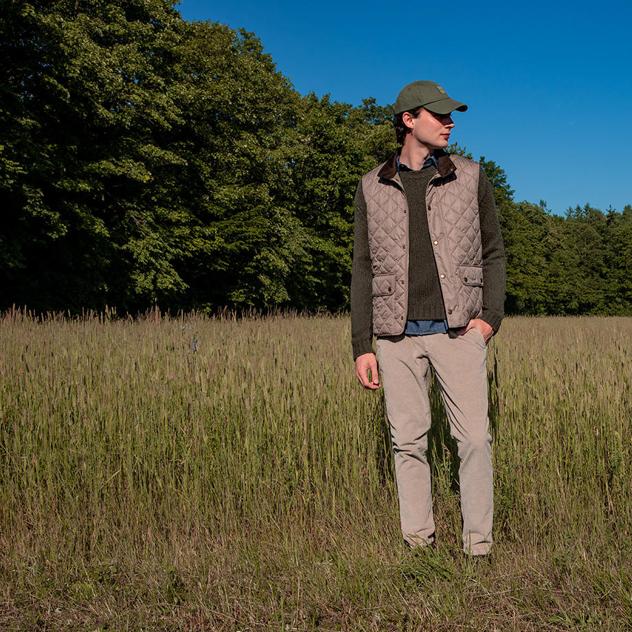 Ein junger Mann in Layering-Look steht in einem Feld vor einem Wald