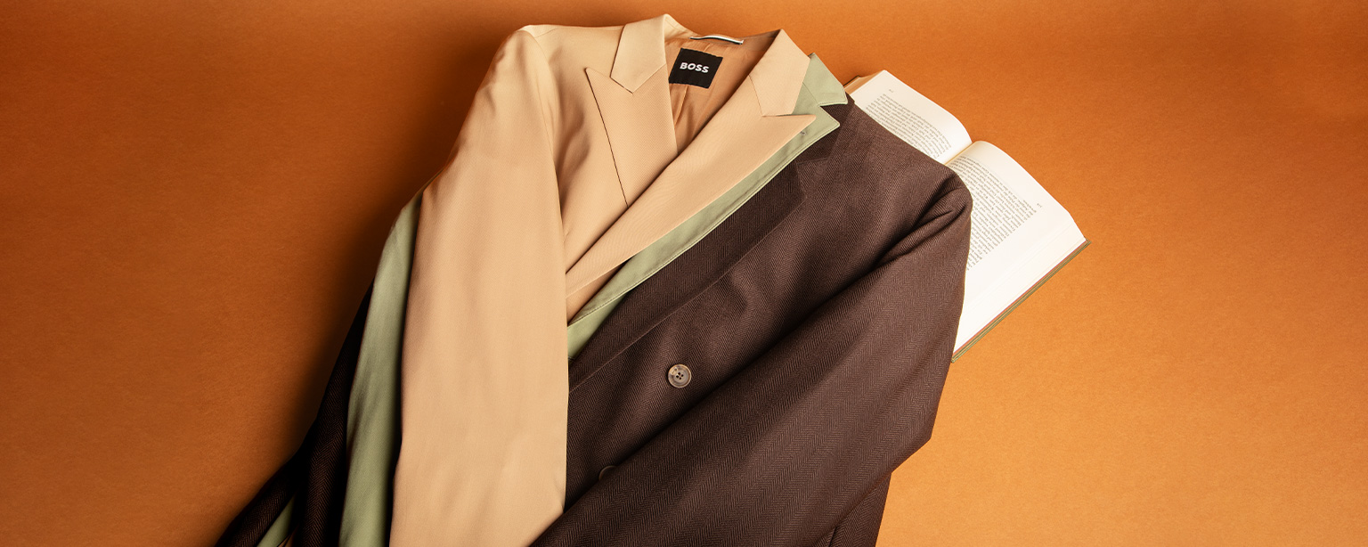 Gefaltete Herren-Sakkos und Jacketts in Beige- und Brauntönen, dekorativ gestapelt auf einem Buch vor orangefarbenem Hintergrund – elegante Business- und Freizeitblazer.