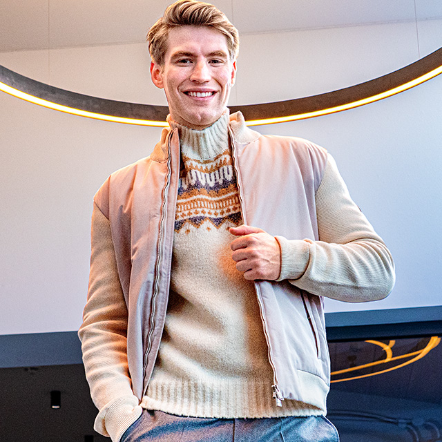 Lächelnder Mann in beigem Norwegerpullover und hellem Blouson unter modernem Lichtring – gemütliche Strickmode für stilvolle Winterlooks bei Herrenausstatter.de.