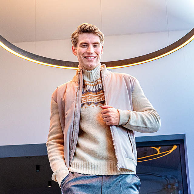 Lächelnder Mann in beigem Norwegerpullover und hellem Blouson unter modernem Lichtring – gemütliche Strickmode für stilvolle Winterlooks bei Herrenausstatter.de.