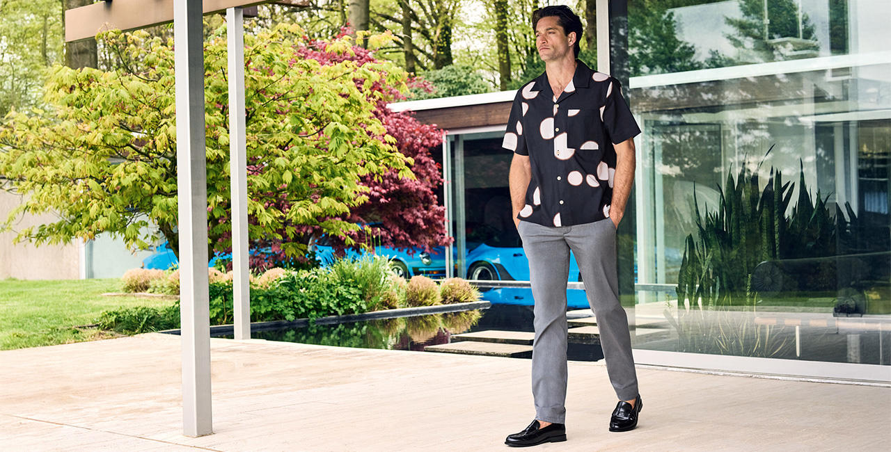 Mann in modernem Sommer-Outfit mit kurzärmligem schwarzem Hemd mit weißen Punkten, grauer Hose und schwarzen Loafern steht entspannt vor einer Glasfassade mit Blick auf Garten und Pool – stilvolle Herrenmode für warme Tage.