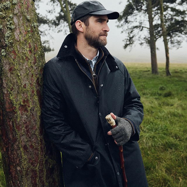 Ein Mann trägt eine Barbour Wachsjacke und lehnt an einem Baum in der Natur