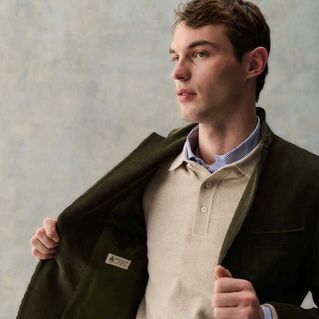 Ein junger Mann in beigem Poloshirt und Barbour Jacke vor einer grauen Wand