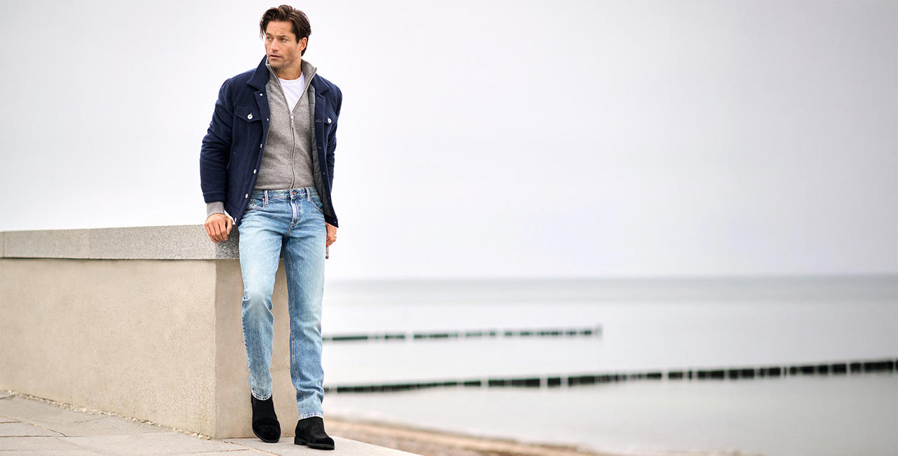 Mann in hellblauer Jeans, dunkelbauer Jacke und grauem Cardigan lehnt an einer Betonbrüstung mit Meerblick – urbaner Denim-Look für Herren.