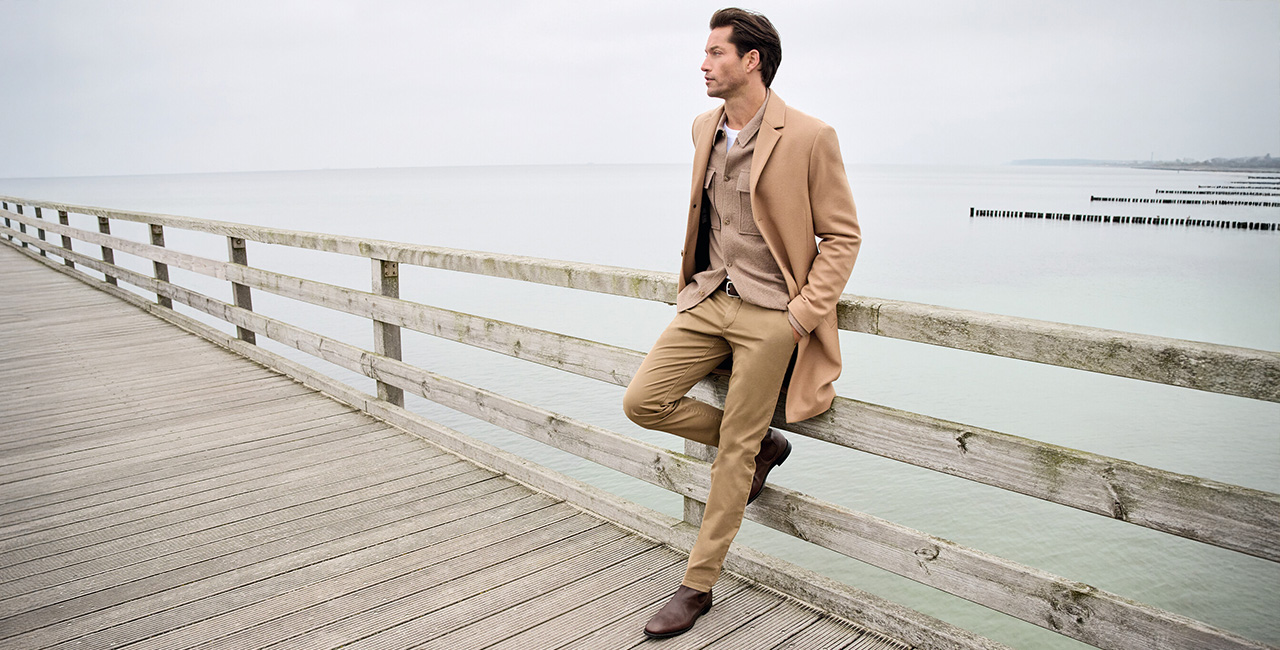 Stilvoller Mann im beigen Hose, Overshirt und Mantel sitzt lässig auf einem Holzsteg am Meer – elegante Hosenmode für Business und Freizeit.