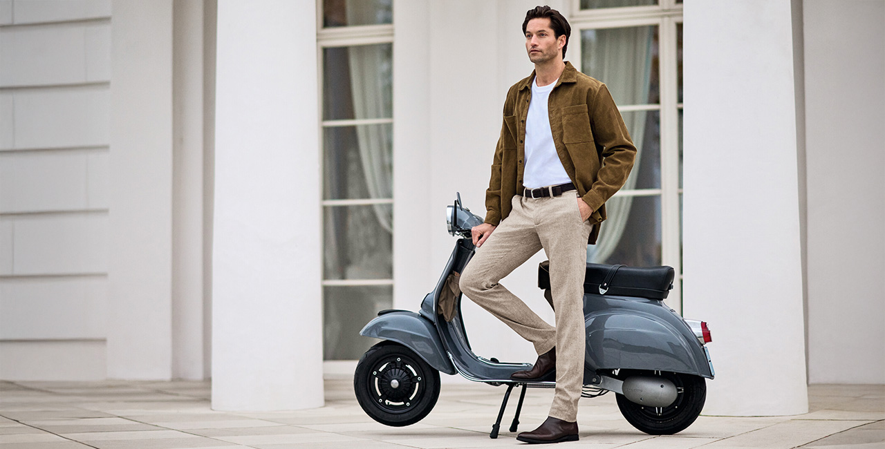 Mann in beiger Chino und brauner Jacke lehnt an grauer Vespa vor moderner Architektur – stilvoller Herrenlook mit Chinohose.