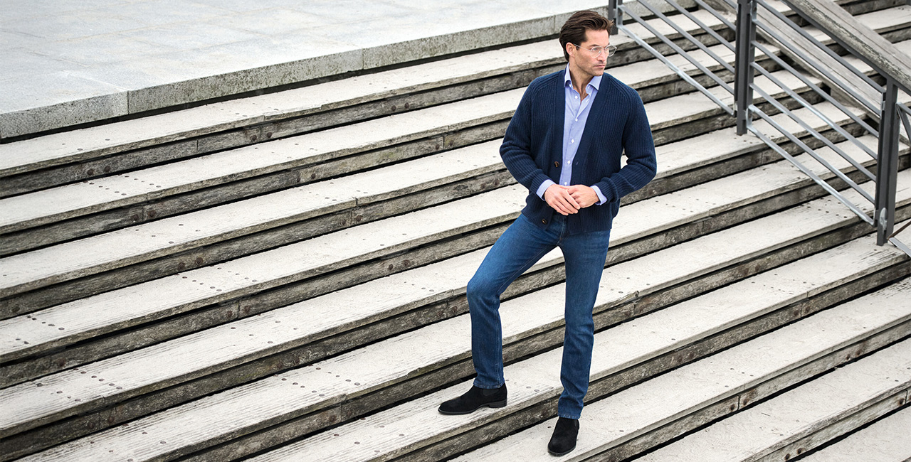 Mann in dunkelblauer Jeans, Strickcardigan und hellblauem Hemd steht auf moderner Steintreppe – Business Casual Herrenmode mit smartem Denim.
