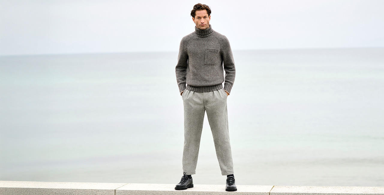 Mann in grauen Hose und Strickpullover steht vor Meerblick – stilvolle und bequeme Hosen für Herren