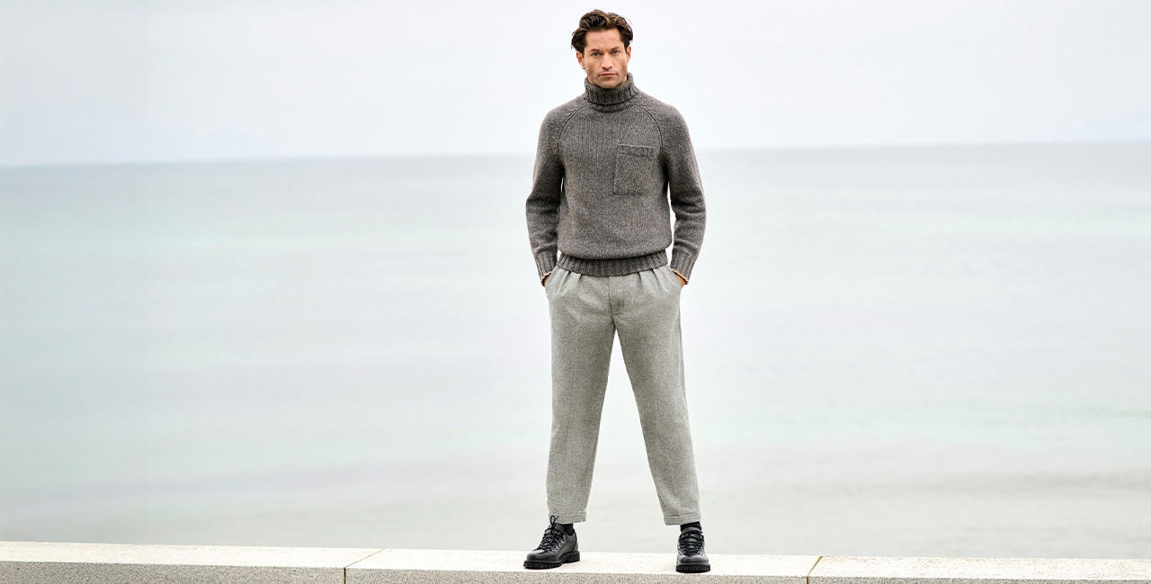Mann in grauen Hose und Strickpullover steht vor Meerblick – stilvolle und bequeme Hosen für Herren