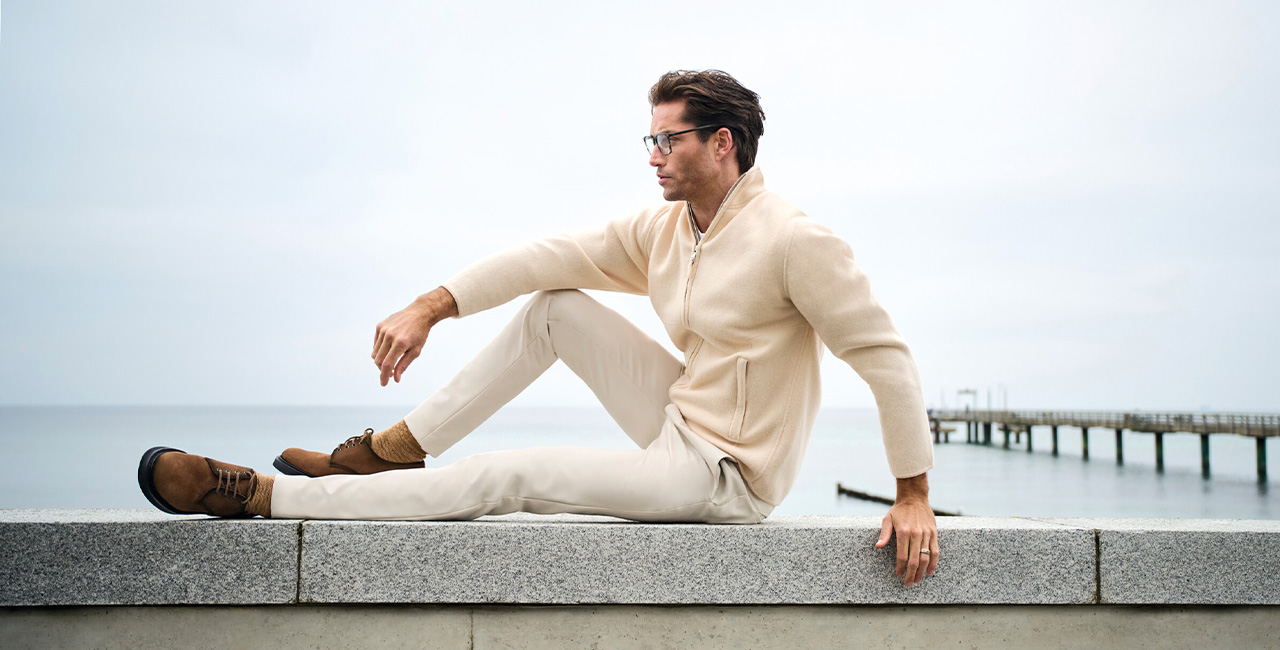 Mann in weißem Strickpullover und heller Hose sitzt entspannt auf einer Mauer mit Meerblick – moderner Freizeitlook in Weiß.
