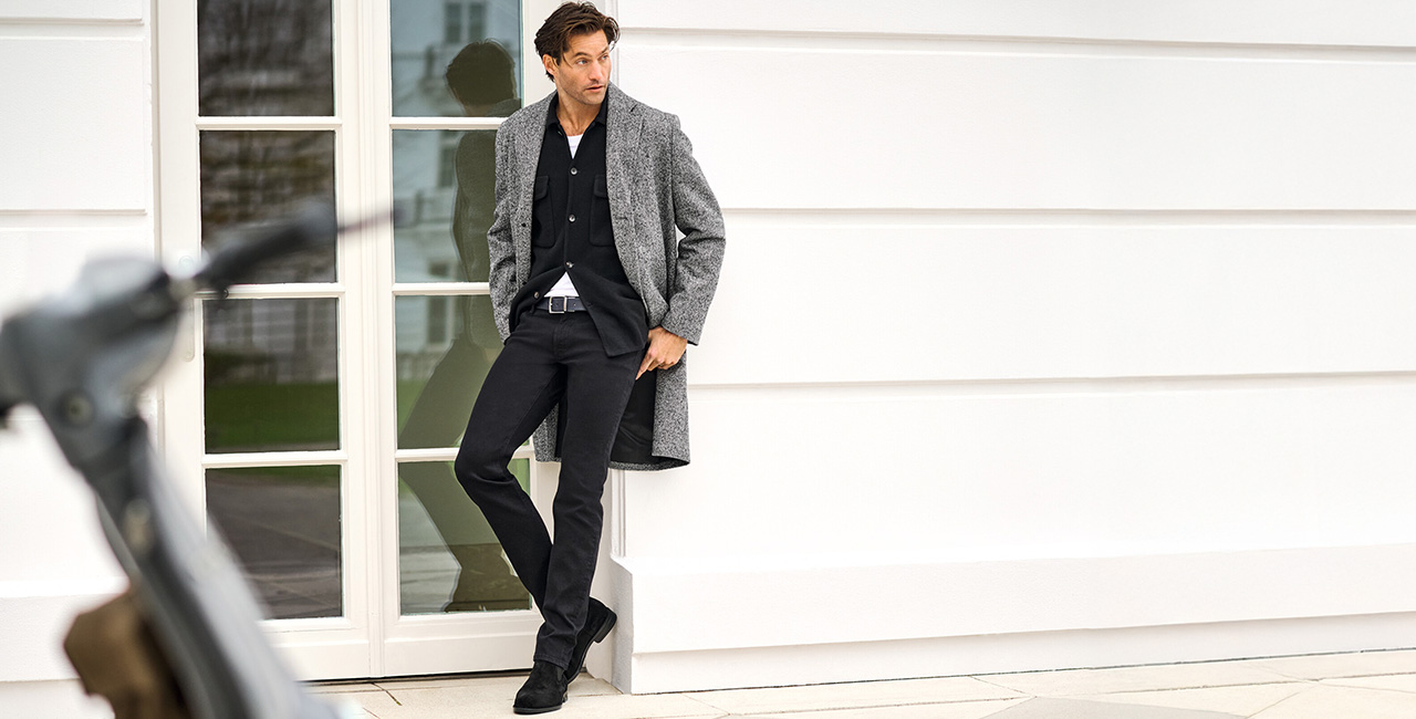 Eleganter gekleideter Mann mit grauem Mantel und schwarzer Hose lehnt entspannt an einer Hausfassade – stilvolle Business-Mode für Herren.