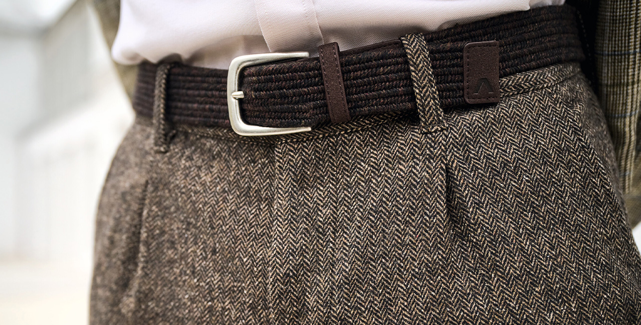 Nahaufnahme eines braunen Stoffgürtels mit Metallschnalle in einer gemusterten Anzughose – elegantes Herrenmode-Accessoire.