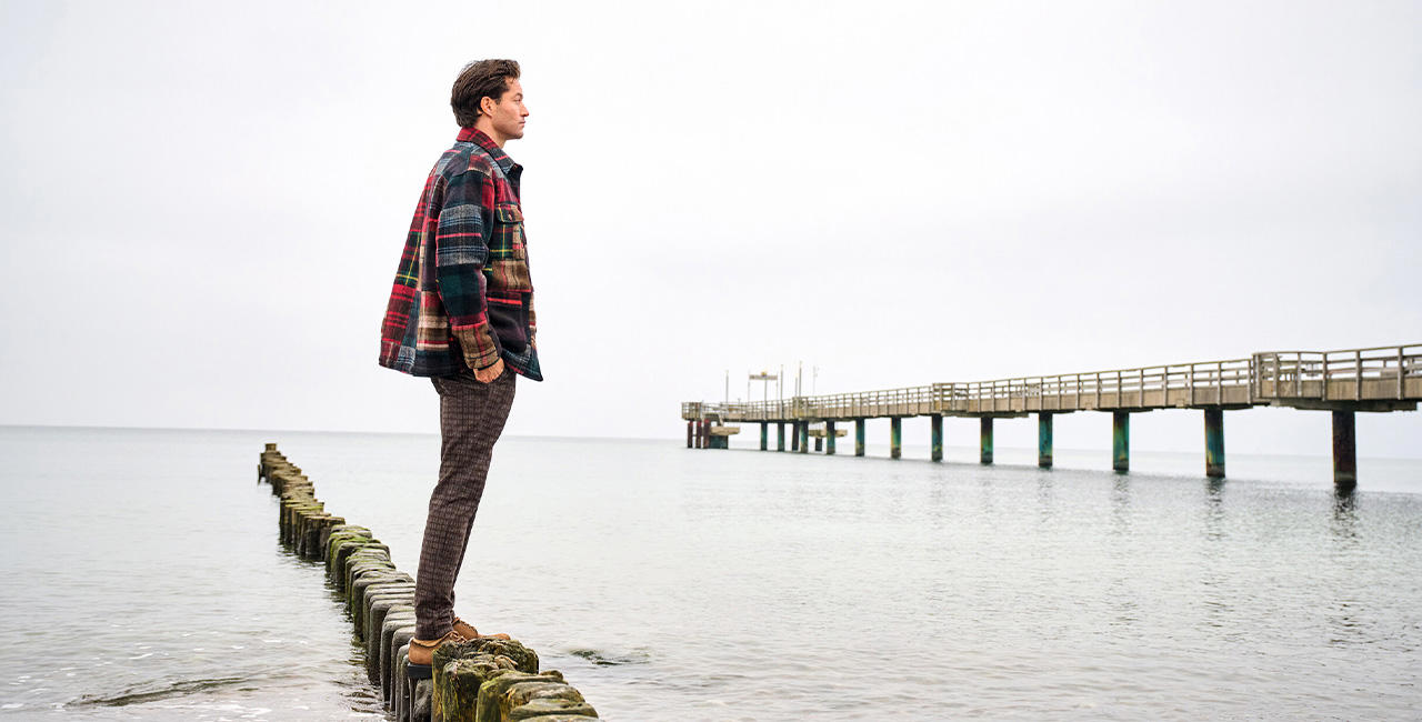 Mann im karierten Overshirt blickt aufs Meer, steht auf Holzpfählen am Strand – lässige Herrenmode für den Herbst.