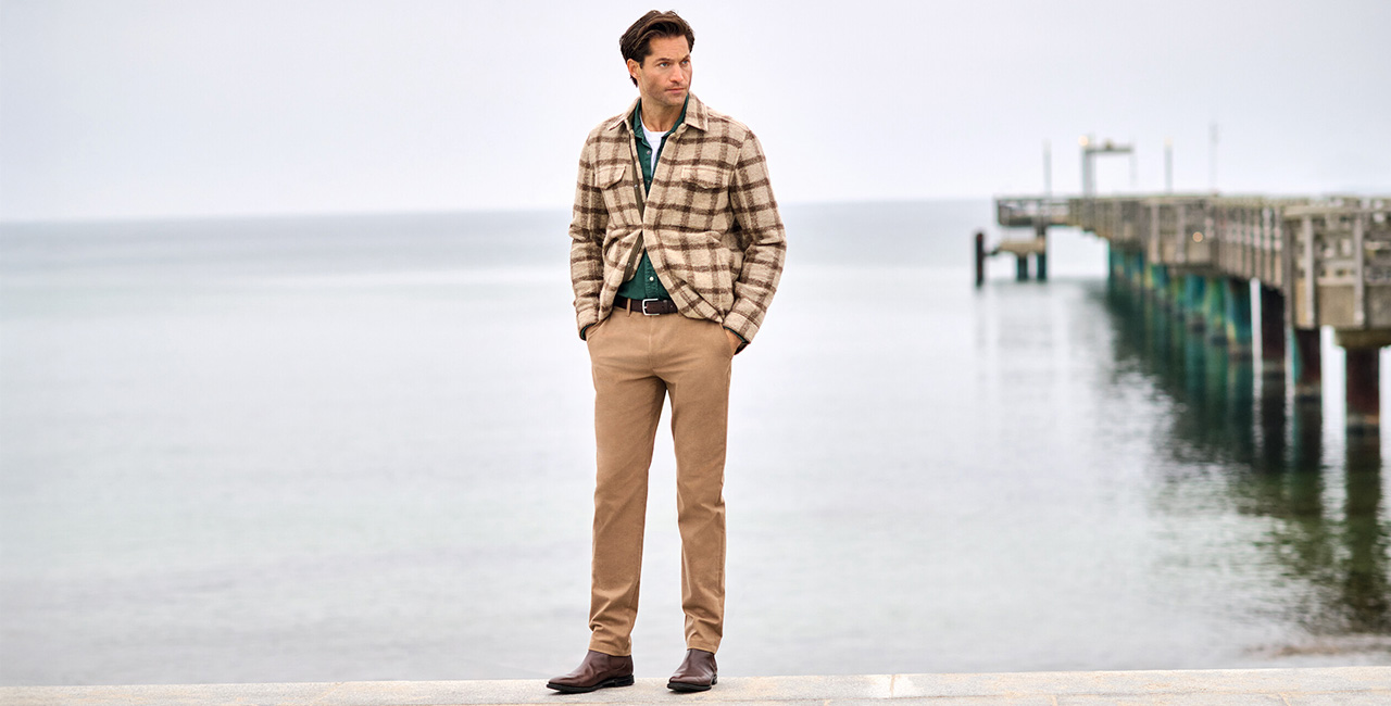 Mann im Smart-Casual-Look mit kariertem Sakko, Hemd und Chinohose steht am Meer. Im Hintergrund ist ein Steg zu sehen – stilvolle Herrenmode für Freizeit und Büro