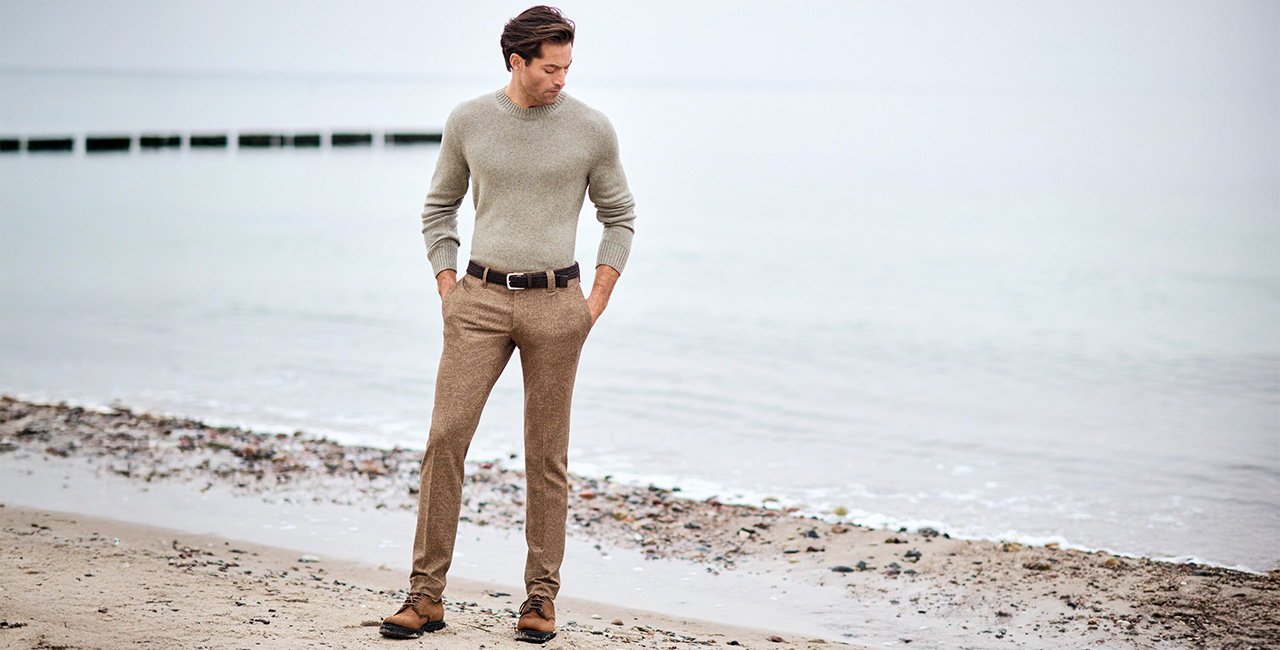 Elegant gekleideter Mann in beigem Strickpullover und brauner Bügelfaltenhose steht am Strand – stilvoller Dressy-Look für Herrenmode.