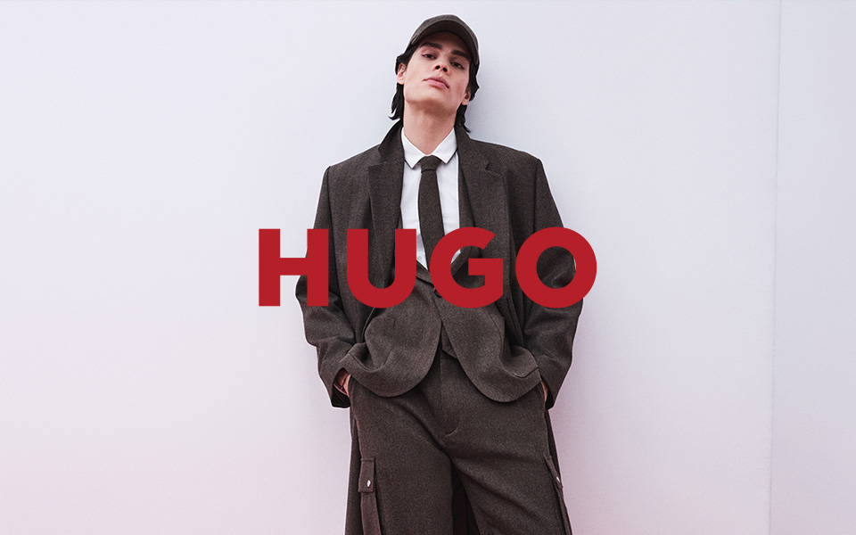 Ein Mann in einem braunen Anzug lehnt an einer weißen Wand. Mittiges Logo von Hugo