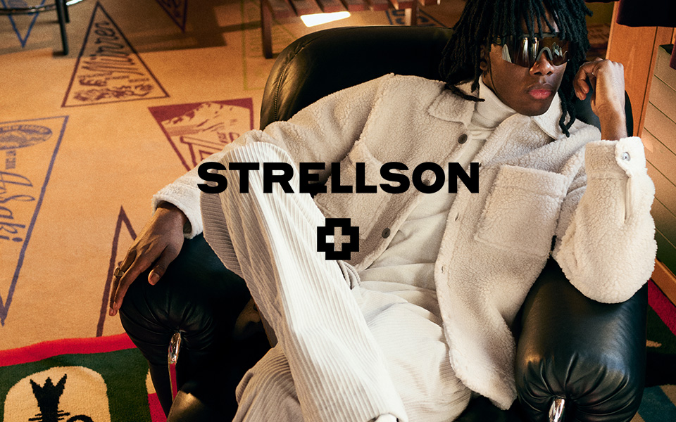 Ein junger Mann in einem cremefarbenen Outfit sitzt auf einem Ledersessel. Mittiges Logo von Strellson
