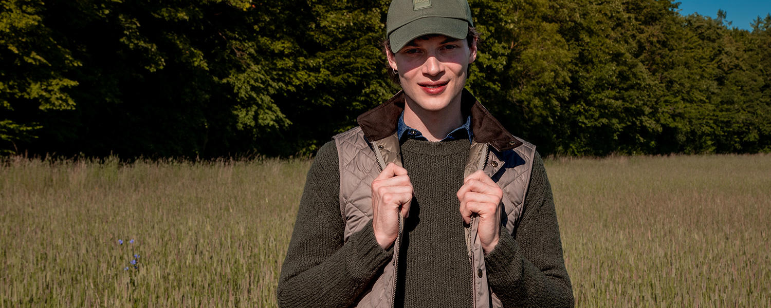 Ein junger Mann steht in einem Kornfeld vor einem Wald. Er trägt einen Layering-Look bestehend aus einem Jeanshemd, einem Pullover und einer Weste.