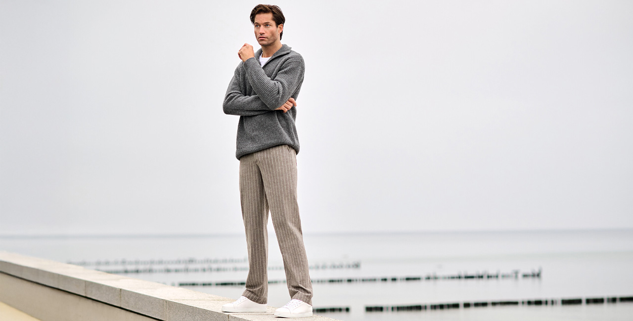 Mann in grauem Strickpullover und brauner Flanellhose mit feinem Streifenmuster steht entspannt am Meer – gemütlicher Herrenmode-Look für kühle Tage.