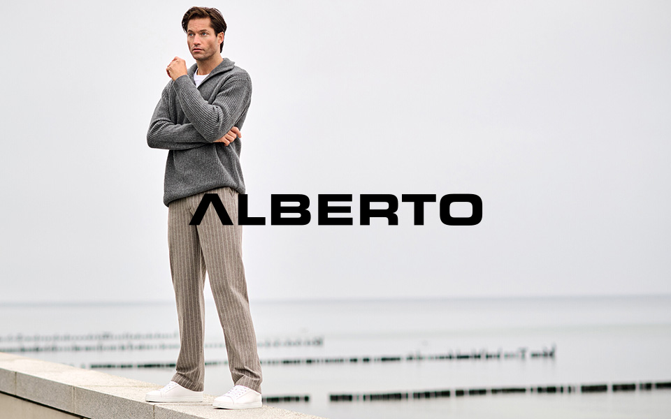 Ein Mann steht am Hafen in einer weiten Hose mit Streifen und einem grauen Cardigan. Mittiges Logo von Alberto