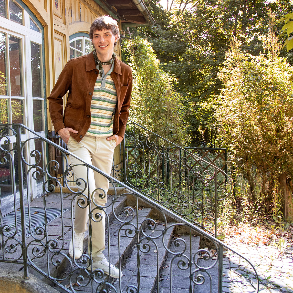 Ein junger Mann steht vor einem alten Haus auf einer Steintreppe. Er trägt ein gestreiftes Polo in herbstlichen Farben sowie eine cognacfarbene Lederjacke
