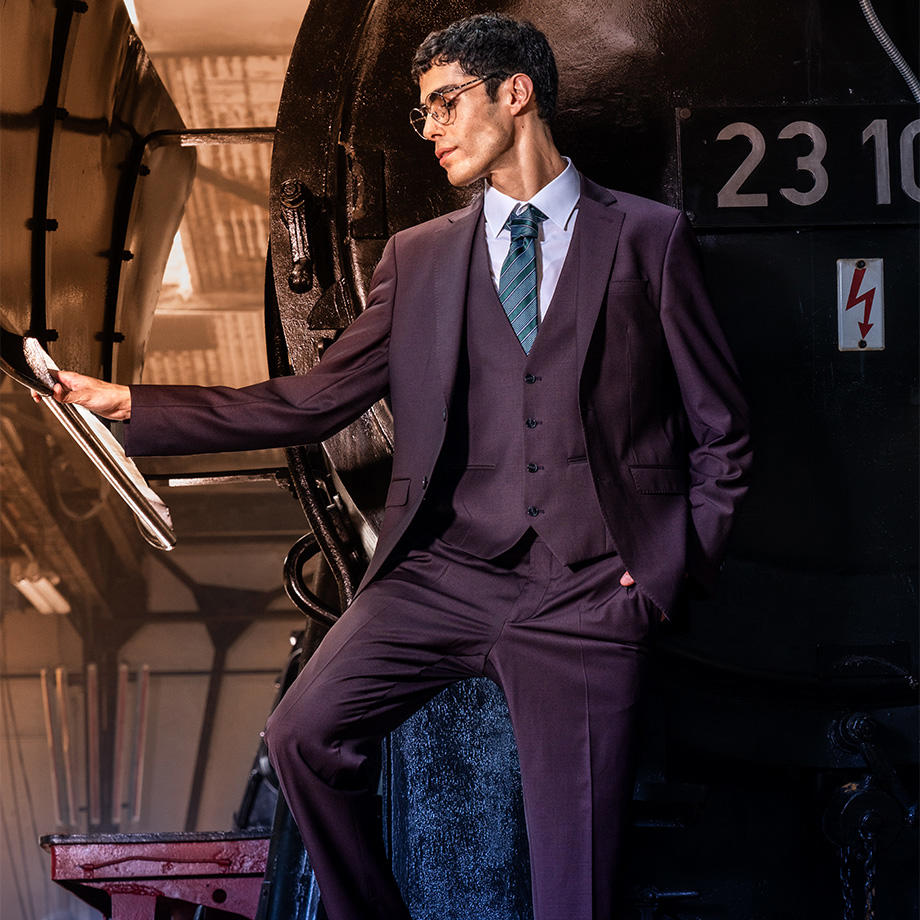 Ein Mann trägt einen dunkelvioletten dreiteiligen Anzug mit Weste und blauer Krawatte, lehnt lässig an einer alten Lokomotive und hält sich mit einer Hand an einem Metallgriff fest.