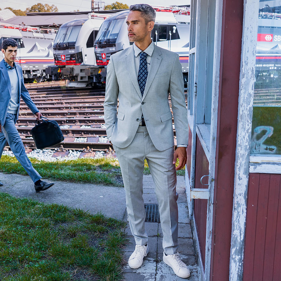 Ein Mann in einem hellgrauen Anzug mit gemusterter Krawatte und weißen Sneakern steht vor einem Gebäude neben Bahngleisen, während im Hintergrund ein weiterer Mann im blauen Anzug mit einer Tasche vorbeigeht.