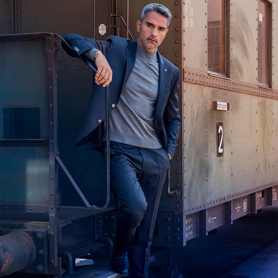 Ein Mann in einem dunkelblauen, dezent gemusterten Anzug mit grauem Rollkragenpullover lehnt lässig an einem alten Zugwaggon und blickt nach vorne.