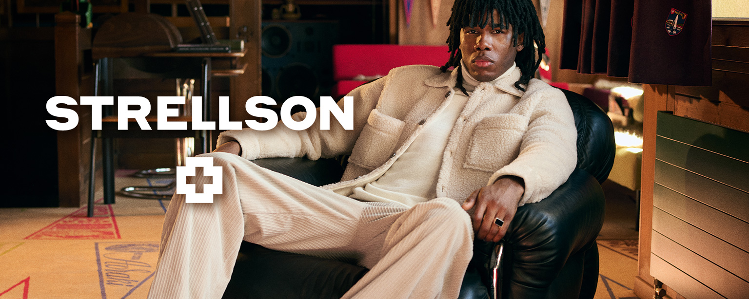 Lässig sitzender Mann im cremefarbenen Cordhose und passender Jacke von Strellson in modernem Wohnzimmer – trendbewusste Herrenmode mit urbanem Charakter.