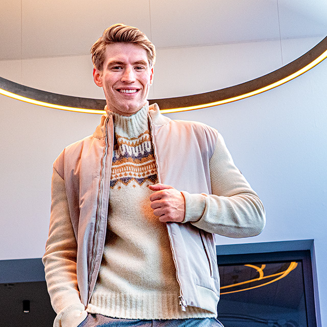 Lächelnder Mann in beigem Norwegerpullover und hellem Blouson unter modernem Lichtring – gemütliche Strickmode für stilvolle Winterlooks bei Herrenausstatter.de.