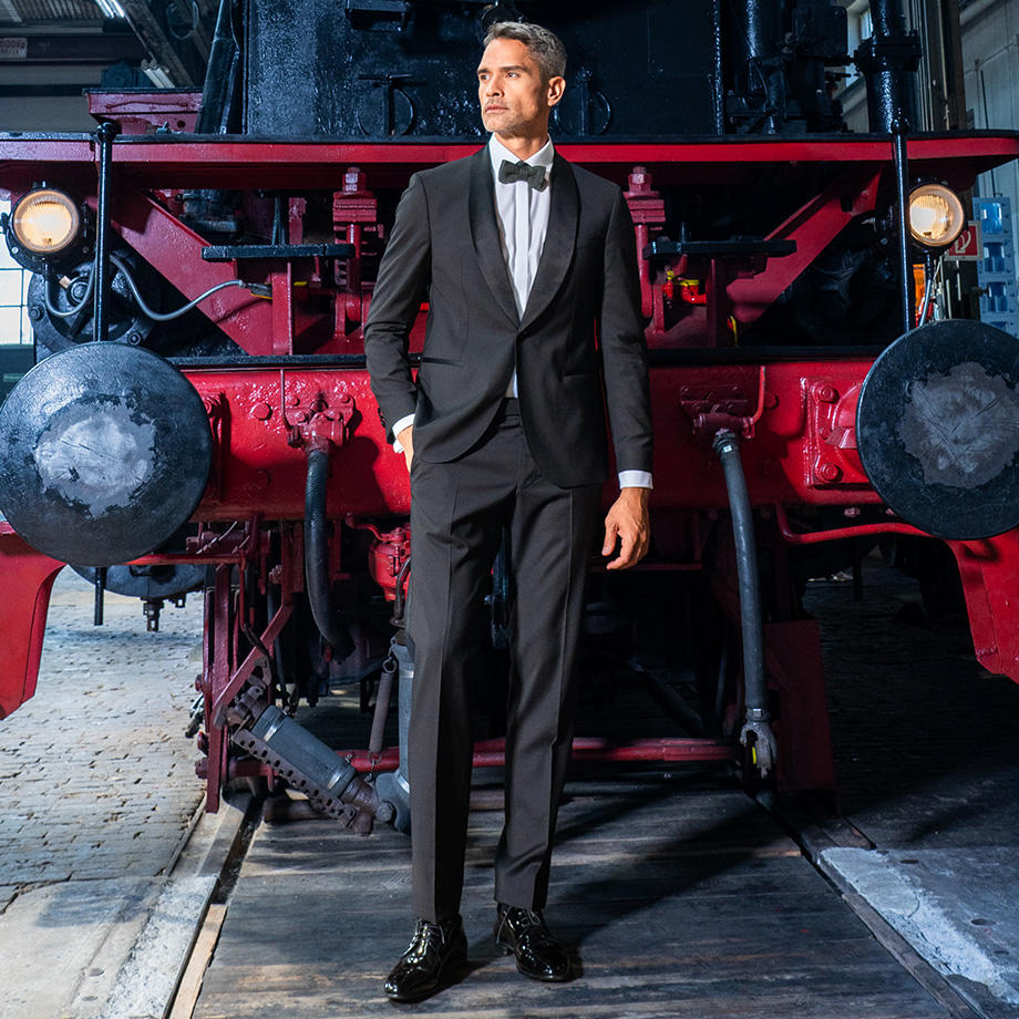 Ein Mann in schwarzem Smoking mit weißem Hemd, schwarzer Fliege und schwarzen Anzugschuhen steht vor einer alten Eisenbahn, hat eine Hand in der Hosentasche und blickt in die Ferne.