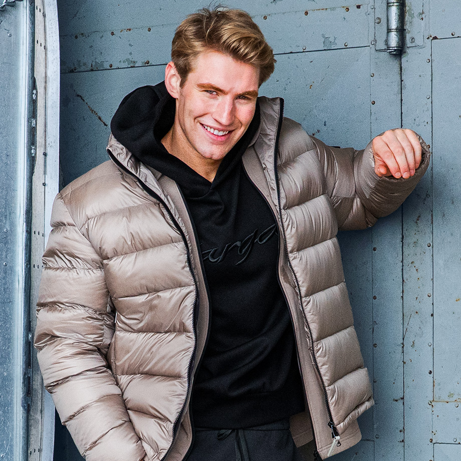 Lächelnder Mann in taupefarbener Steppjacke und schwarzem Hoodie vor industrieller Betonwand – lässige Wintermode für Herren mit urbanem Streetstyle.