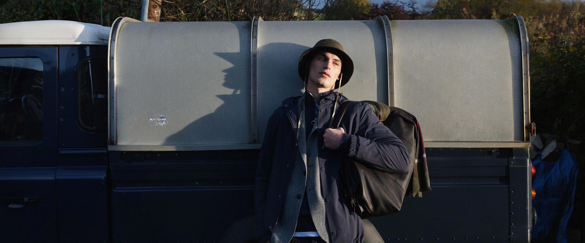 Mann in navyblauer Barbour-Jacke, grauem Schal und Fischerhut lehnt lässig an einem Land Rover – authentischer Outdoor-Look aus der Barbour Tartans Kollektion.