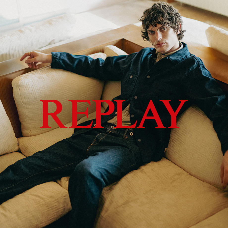 Ein Markenbild von der Marke "Replay". Ein Mann sitzt lässig auf einer hellen Couch in einem Raum mit warmer Lichtstimmung. Er trägt ein dunkles Denim-All-Over-Outfit mit einem hellen Hemd darunter. Sonnenlicht fällt sanft durch das Fenster und unterstreicht den entspannten, stilvollen Replay-Look.