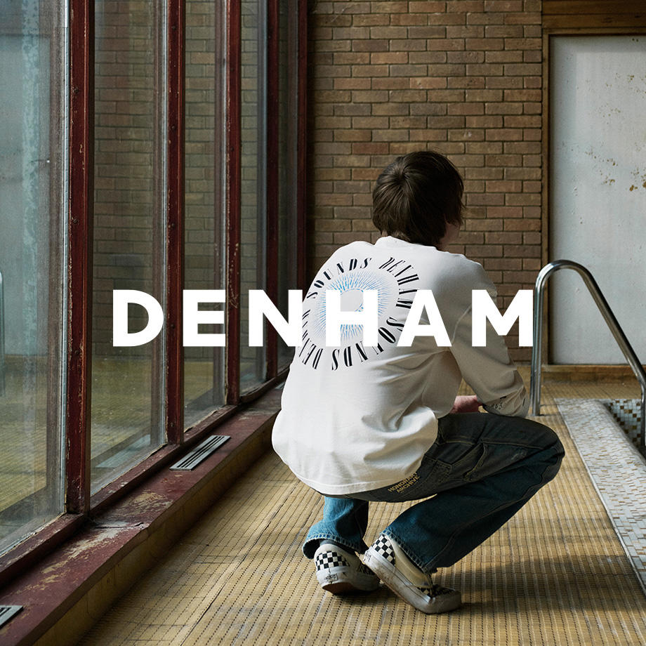 Ein Markenbild von der Marke "Denham". Ein junger Mann hockt am Rand eines leeren, alten Schwimmbeckens mit Backsteinwand und großen Fenstern. Er trägt ein weißes Longsleeve mit Rückenprint, weite Jeans und karierte Slip-On-Sneaker – ein lässiger, urbaner Look in industrieller Umgebung.